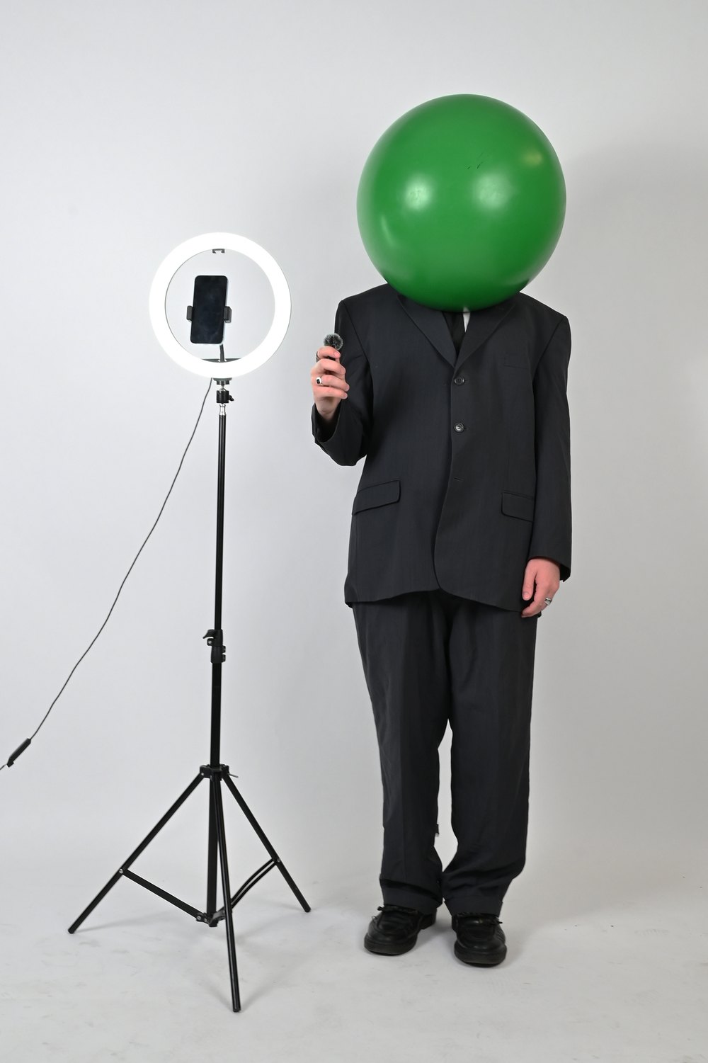 The Goon Guide standing beside the rig: a dark suit, a green inflatable ball worn as a head, a ring light on a tripod holding a phone, and a lapel mic held in one hand.